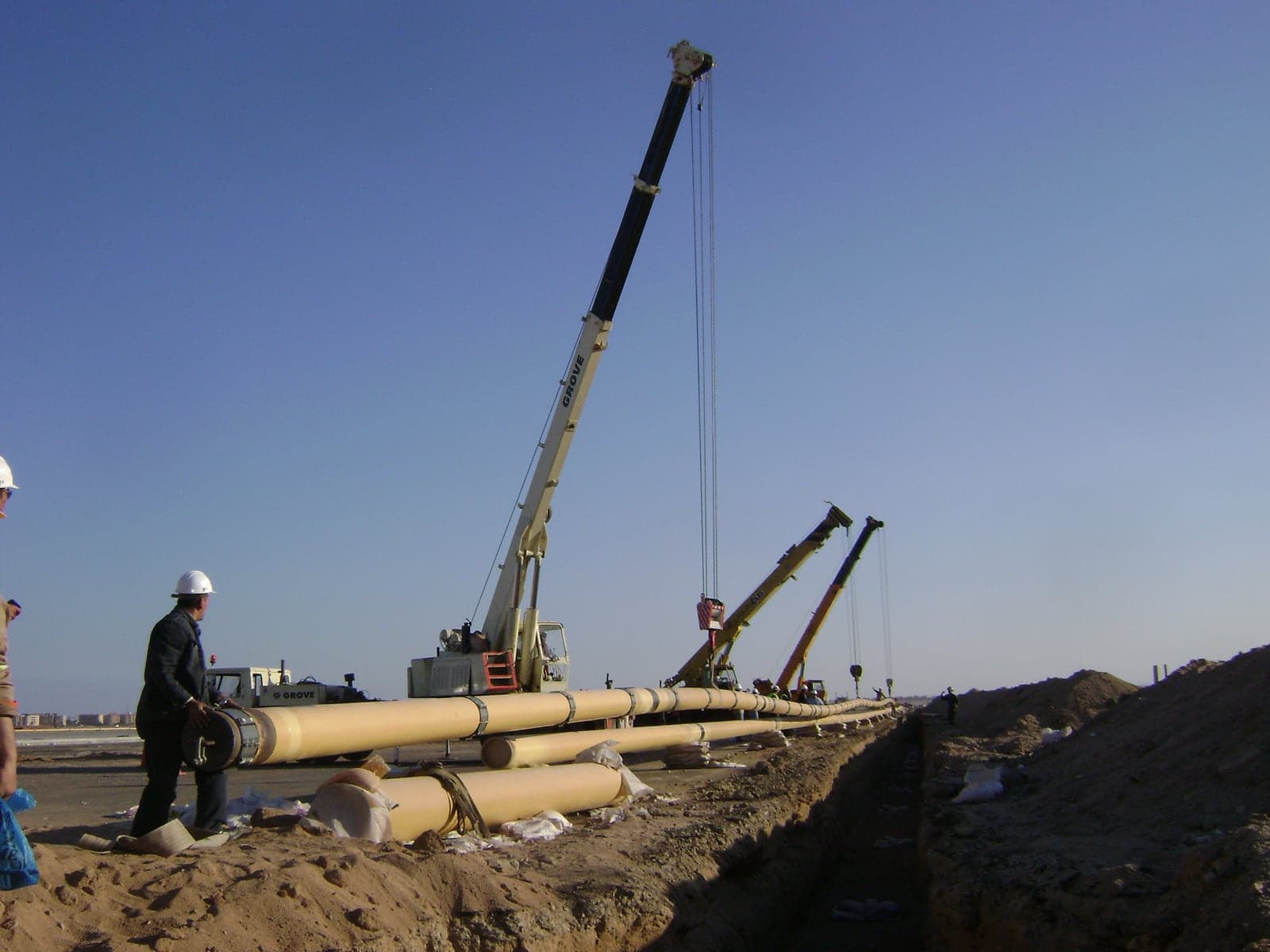 Cranes stringing the 14-inch hydrant mains beside the trench at Hurghada Airport, with the town on the horizon