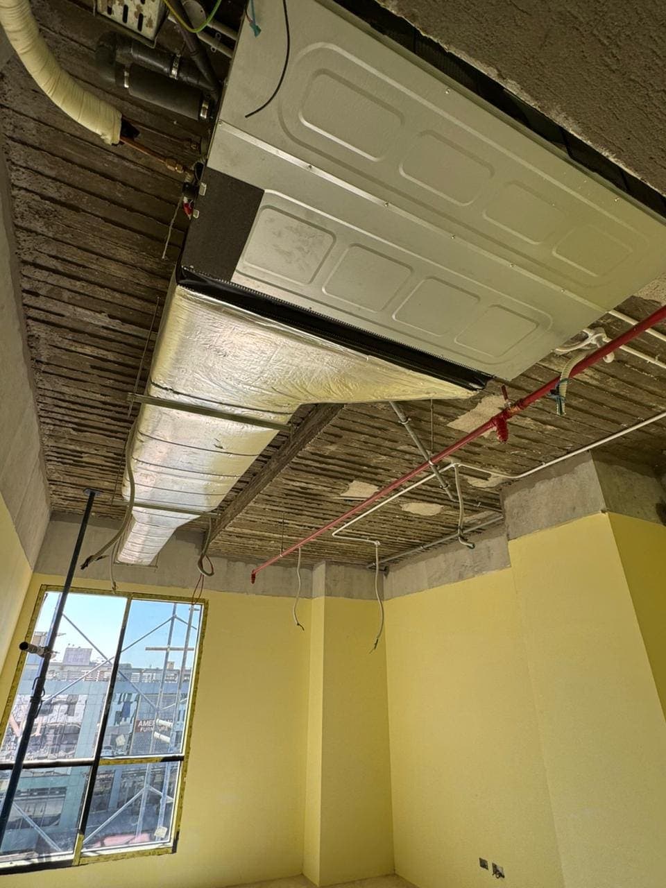 A ducted fan-coil unit and insulated ductwork at soffit level in a bare unit, with a red firefighting pipe and conduit alongside
