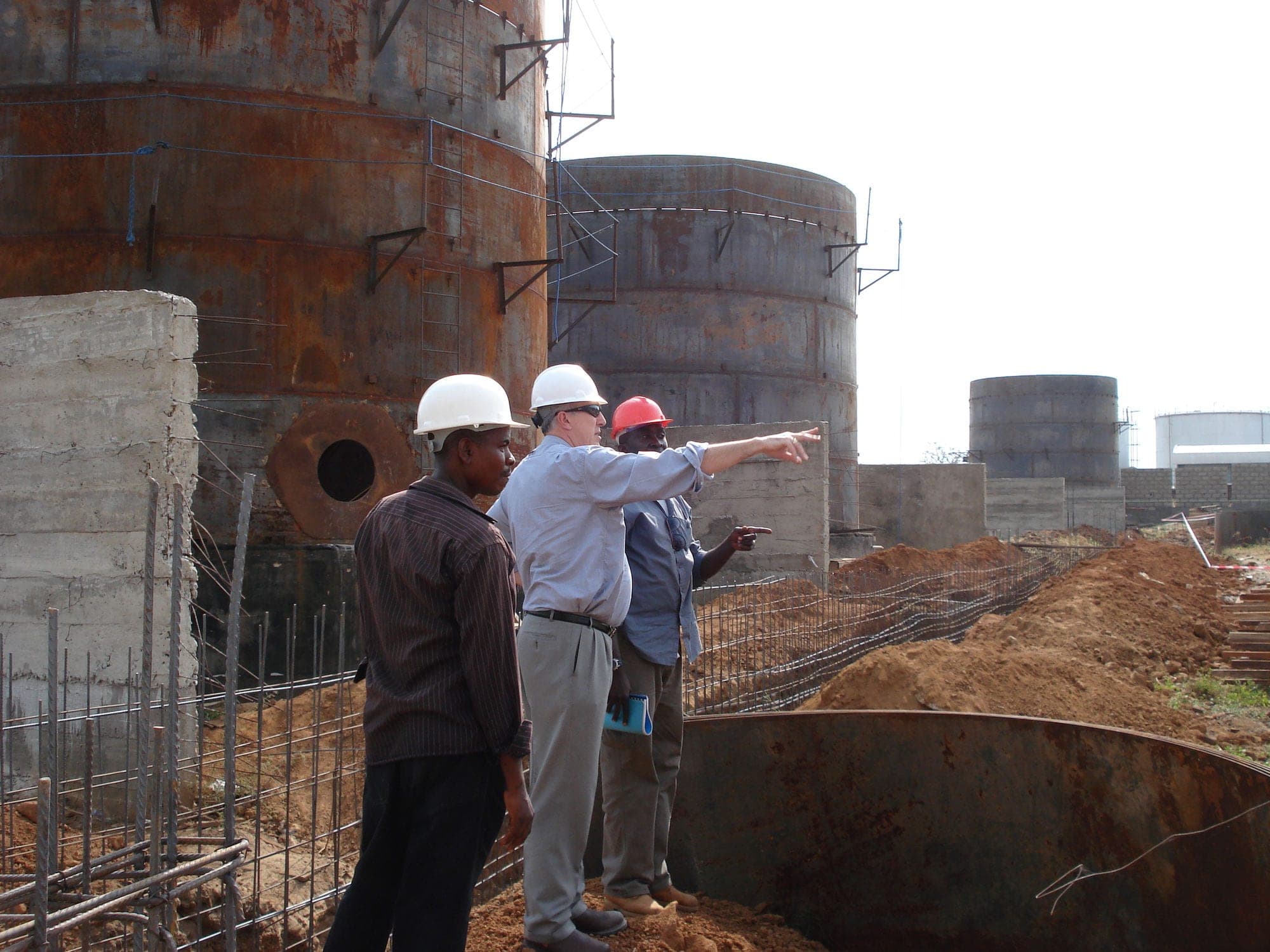 Three engineers on site reviewing the construction of bulk storage tanks at a fuel depot, with tank shells raised and foundations still open