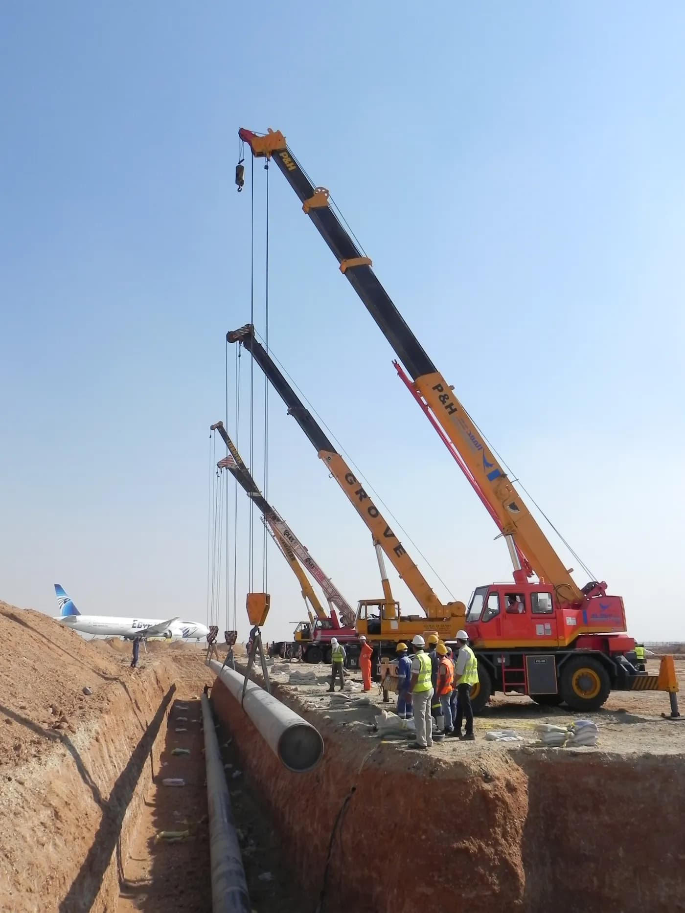 Three mobile cranes lowering a coated jet-fuel hydrant pipeline into a trench at Cairo Airport TB2, an EgyptAir aircraft beyond